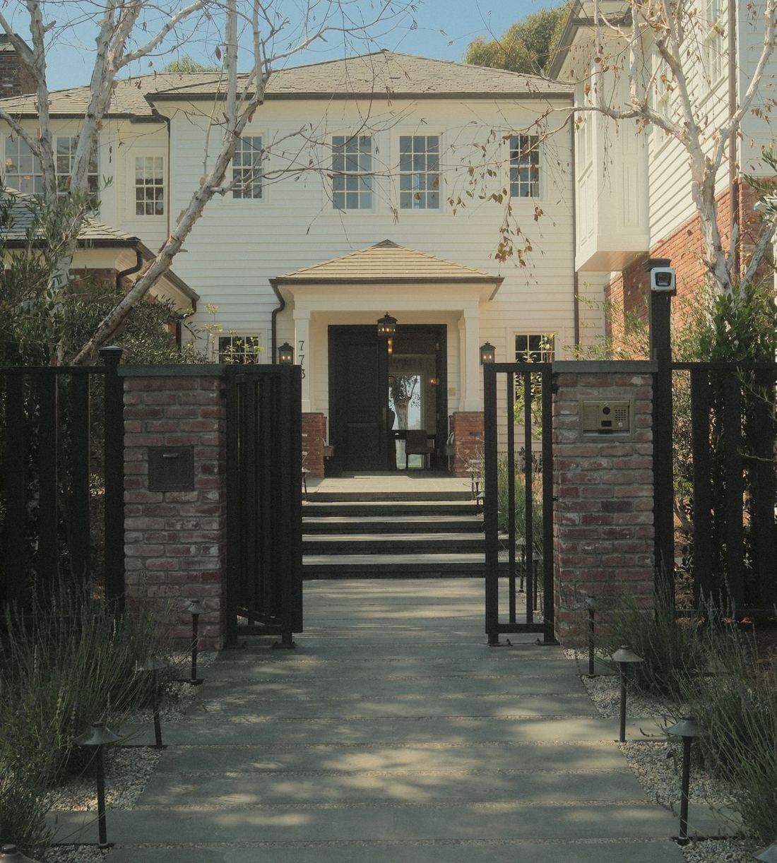 Front entry walkway of bluestone pavers lined with gravel, lavender and path lights, leading through brick pillars and black iron gates to a white two-story home