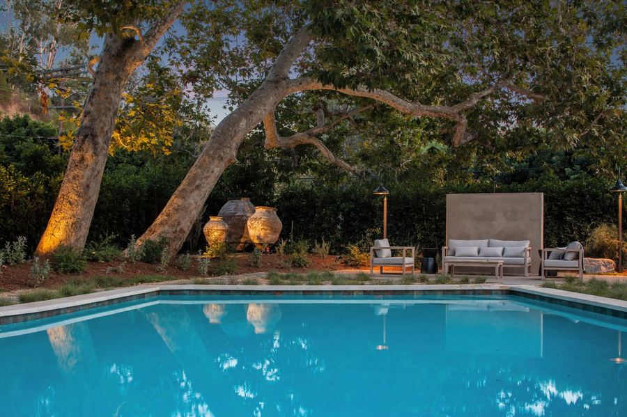 Pool at dusk with uplit sycamore trees, large clay urns and a lounge area against a plaster wall