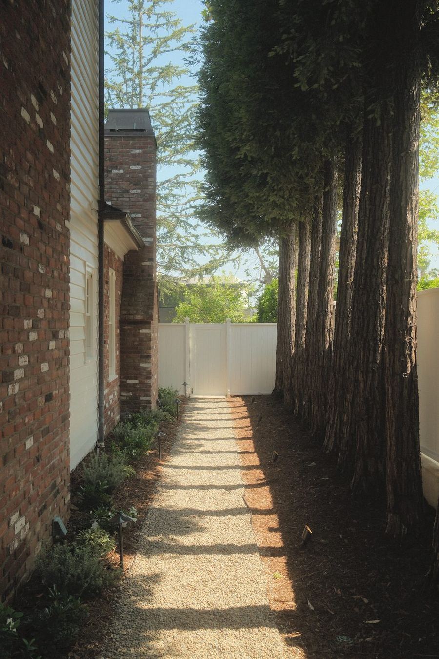 Gravel side-yard path lined with low plantings and path lights between a brick home and tall trees