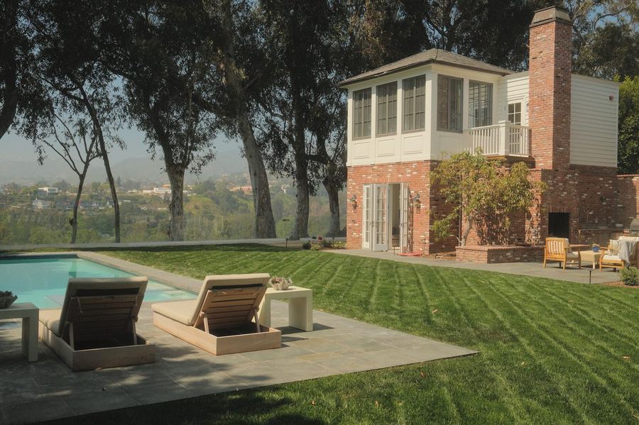 Striped lawn and bluestone pool deck with loungers beside a brick pool house with an outdoor fireplace